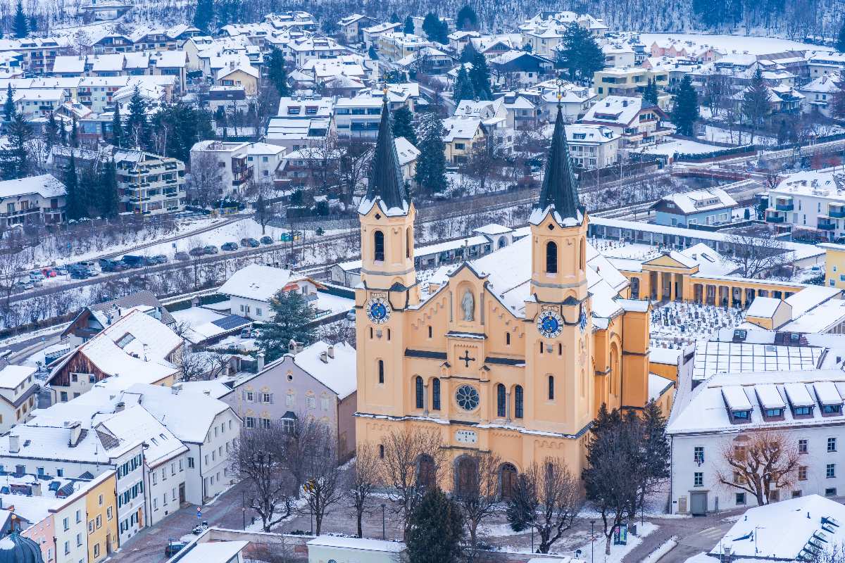 Vista aerea di Brunico innevata con la chiesa di Santa Maria Assunta in primo piano.