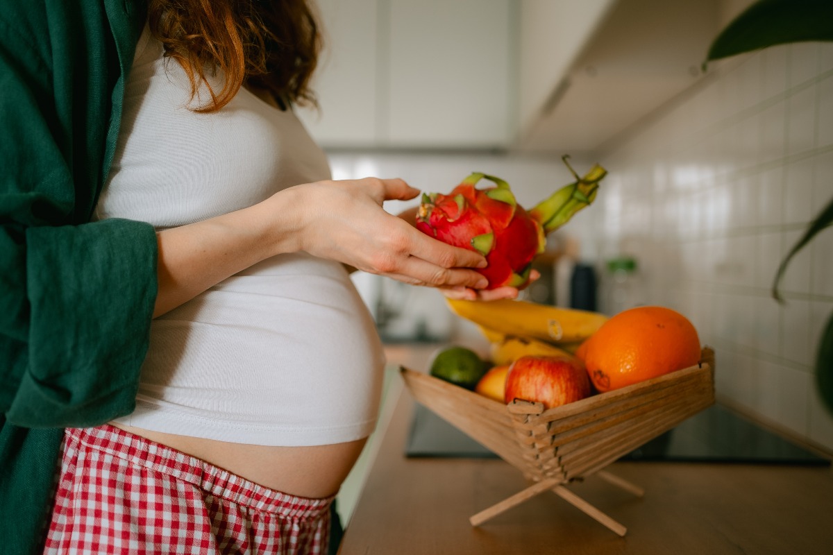 Donna incinta in cucina davanti a un cesto di frutta fresca.